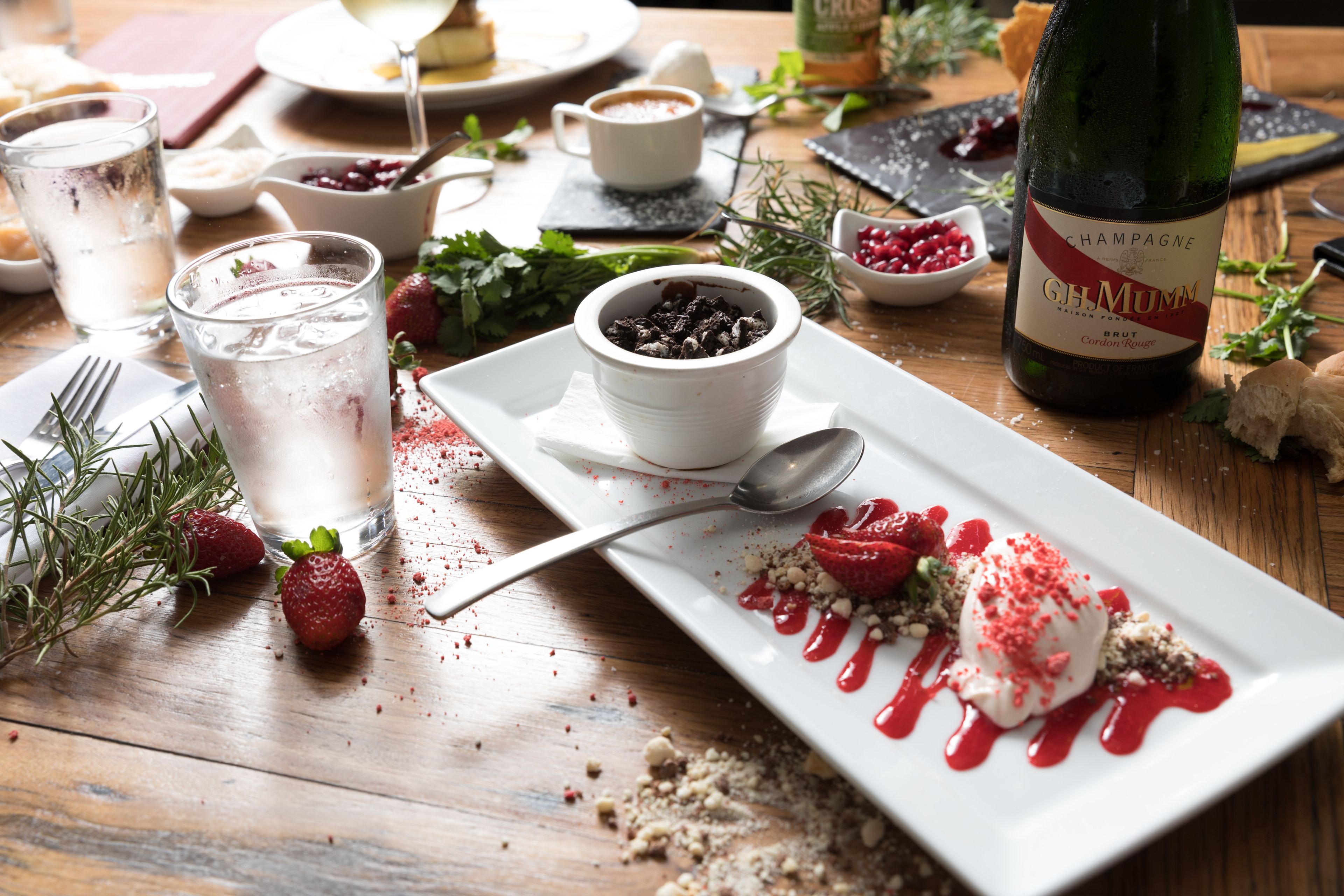 A dessert plate with strawberries and a scoop of ice cream, a bottle of champagne, and a bowl of berries on a wooden table.