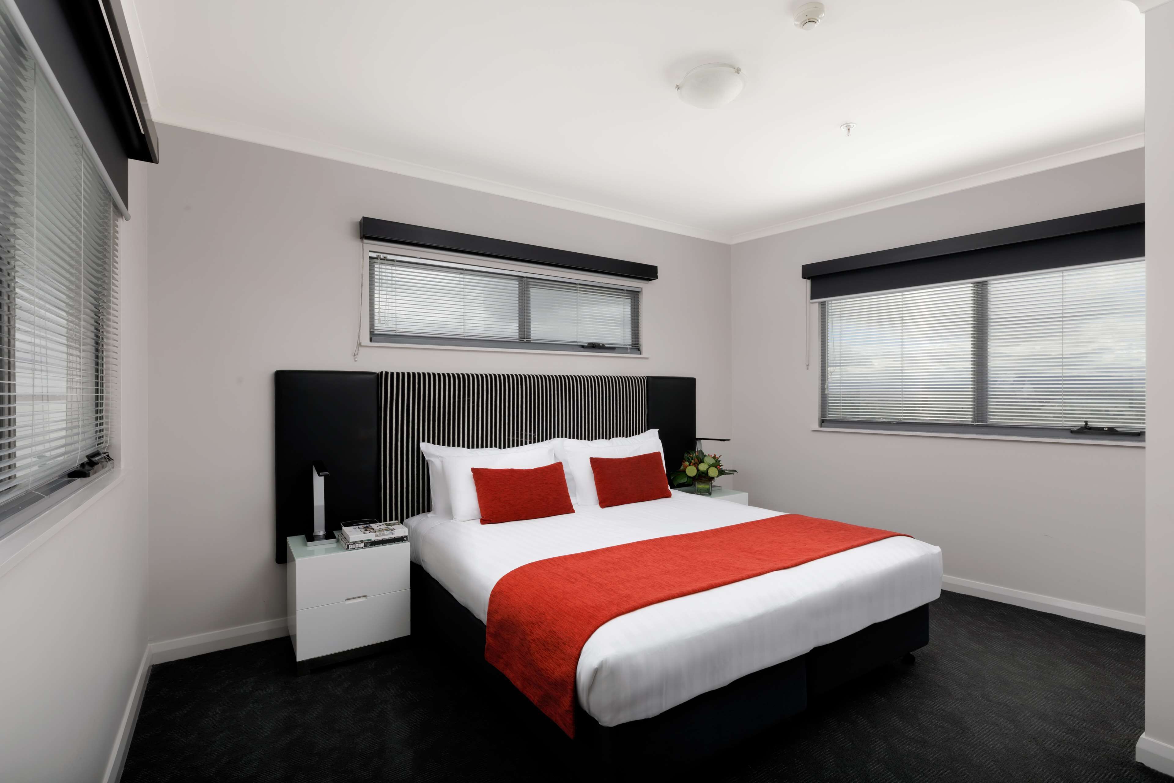 A modern bedroom with a large bed, white bedding, red pillows, and black headboard. There are two bedside tables with lamps, a window with blinds, and a small plant on the nightstand.