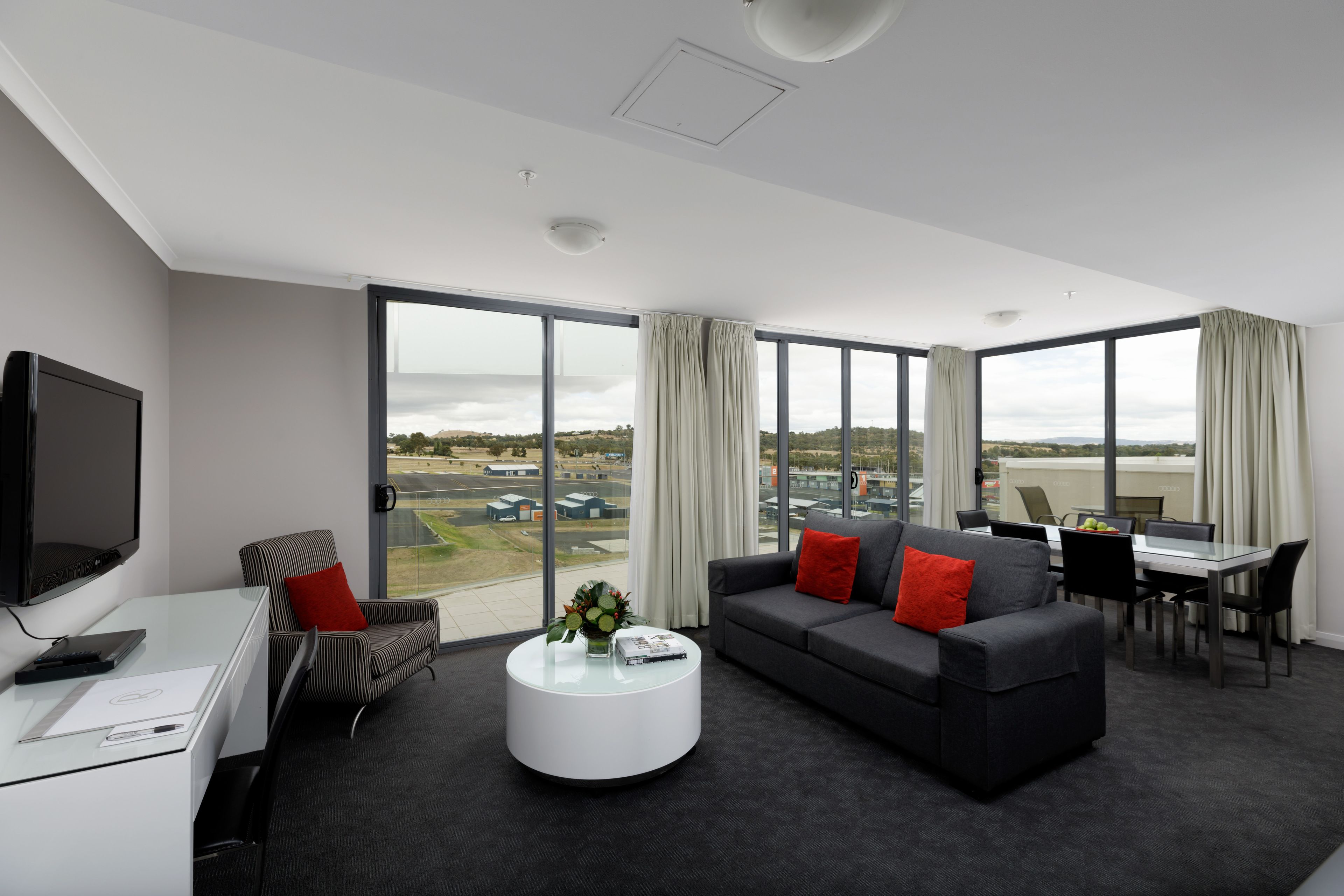 A modern hotel room with a large window, a flat-screen TV, a sofa, a dining table, and a coffee table.