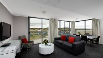 A modern hotel room with a large window, a flat-screen TV, a sofa, a dining table, and a coffee table.