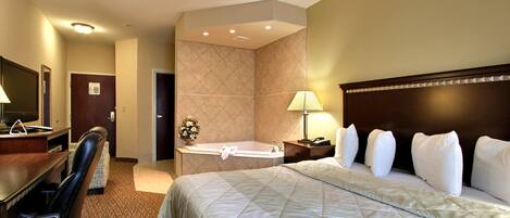 A hotel room with a large bed, a desk with a chair, a desk lamp, a telephone, and a bathroom with a bathtub and a mirror.