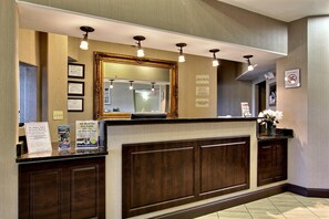 A hotel reception area with a wooden reception desk, framed certificates on the wall, and a large mirror above the desk.