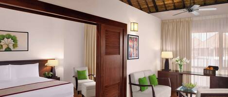 Habitación de hotel con cama, dos sillones, una mesa de comedor, un ventilador de techo y un amplio ventanal con cortinas.
