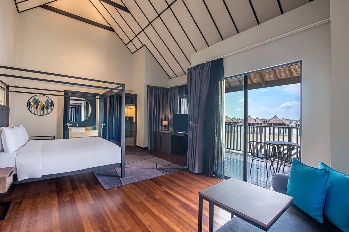 A modern hotel room with a large bed, a balcony with outdoor seating, and a view of the sky.