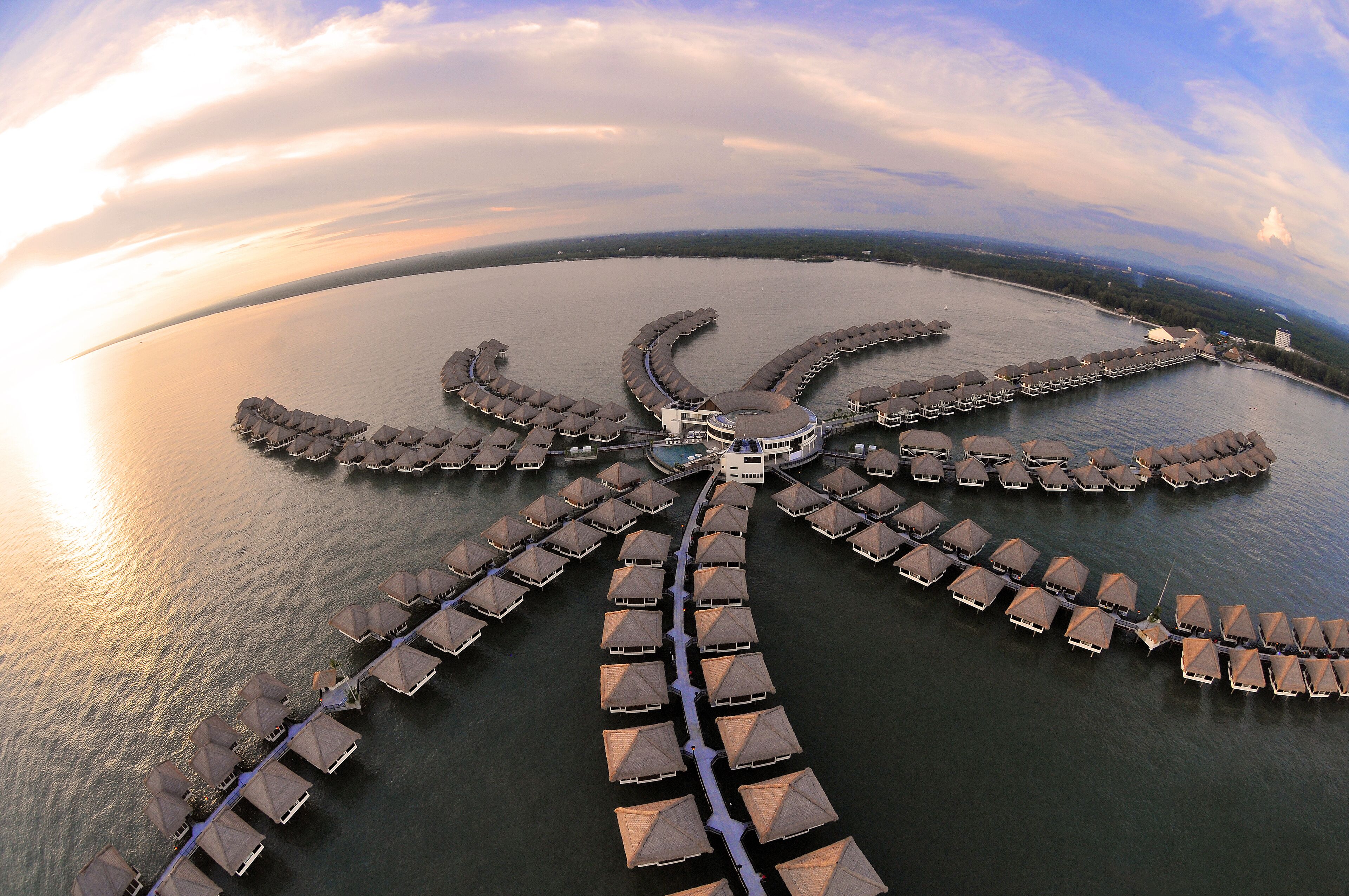 Eine Luftaufnahme eines Resorts mit einem zentralen Gebäude, umgeben von einer kreisförmigen Anordnung von überwasserbungalows, die durch Stege verbunden sind.