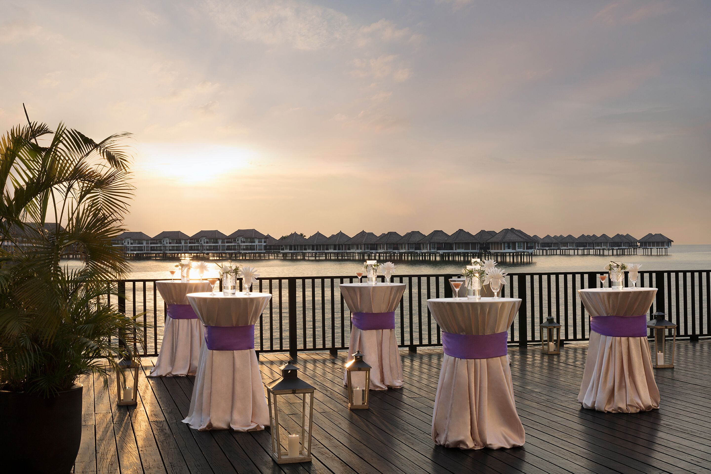 A deck with tables set for an event, overlooking a body of water and a row of overwater bungalows.
