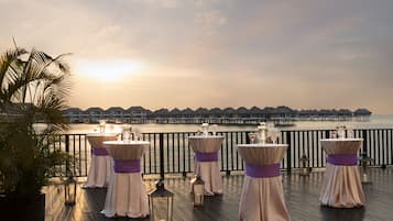 A deck with tables set for an event, overlooking a body of water and a row of overwater bungalows.