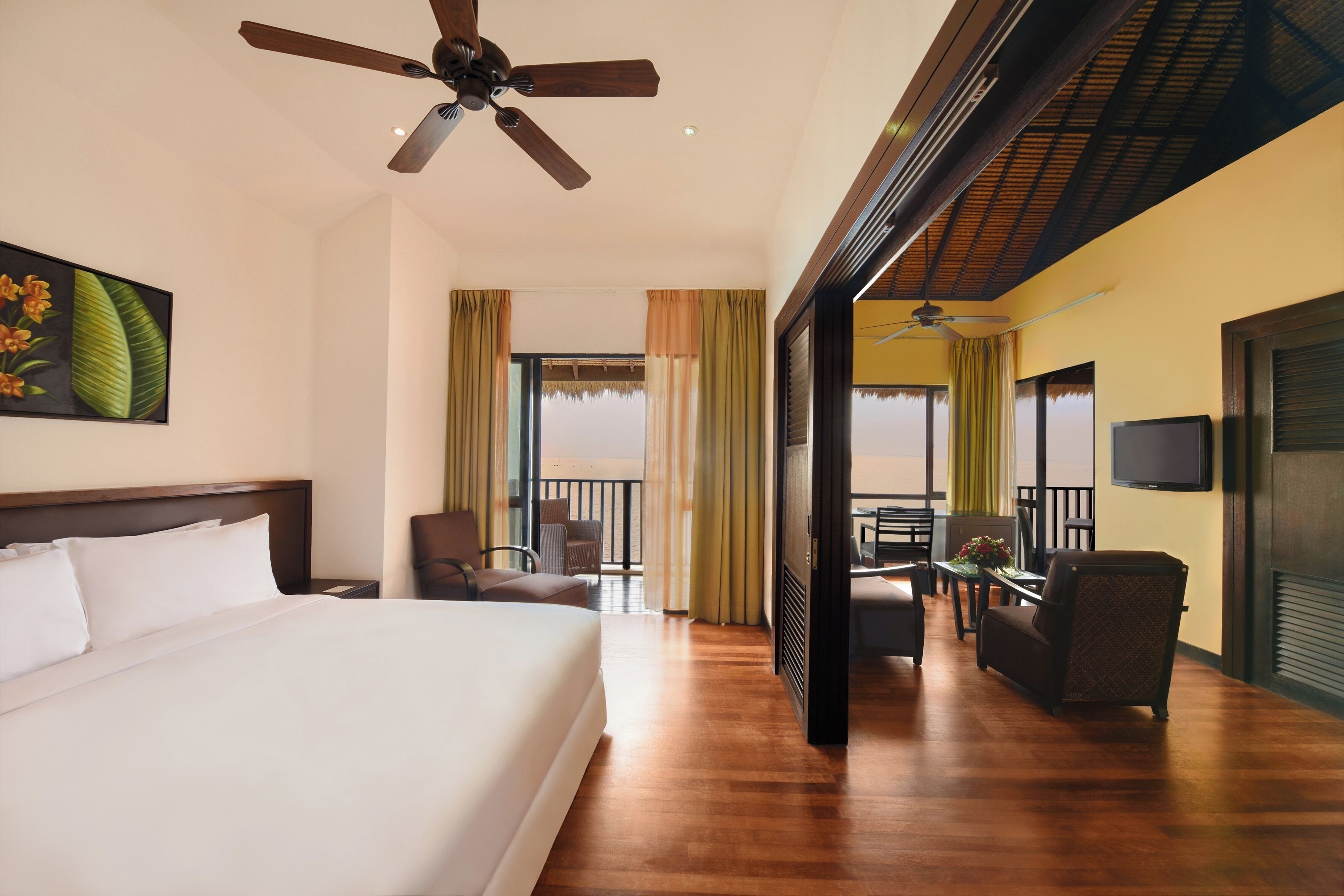 A hotel room with a large bed, a ceiling fan, a balcony with chairs, and a television.