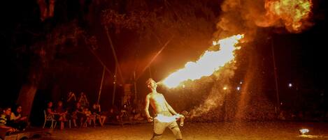 A person performing a fire act on a sandy beach with an audience in the background.