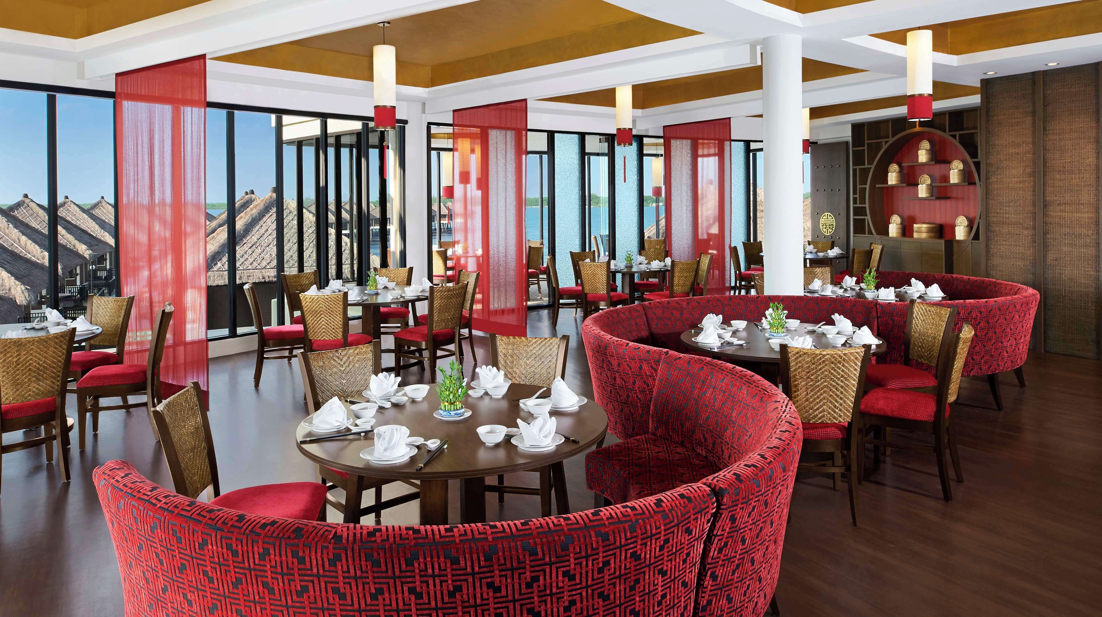 Een restaurant met ronde tafels en rode beklede stoelen, grote ramen met uitzicht op de bergen, en een traditioneel Aziatisch interieur.