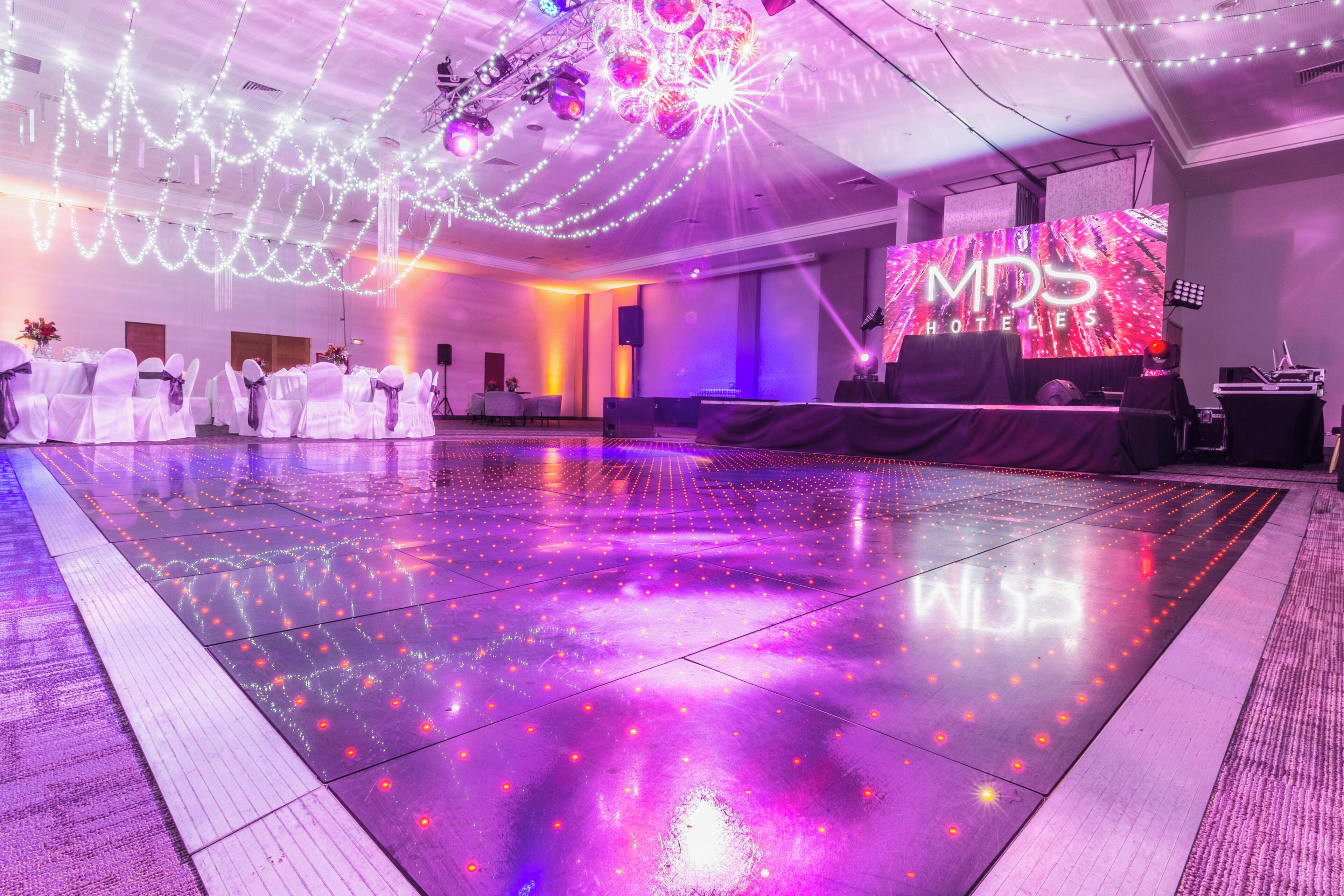 Uma sala de baile de hotel com piso espelhado, palco com sistema de som e uma grande tela LED exibindo o nome do hotel.