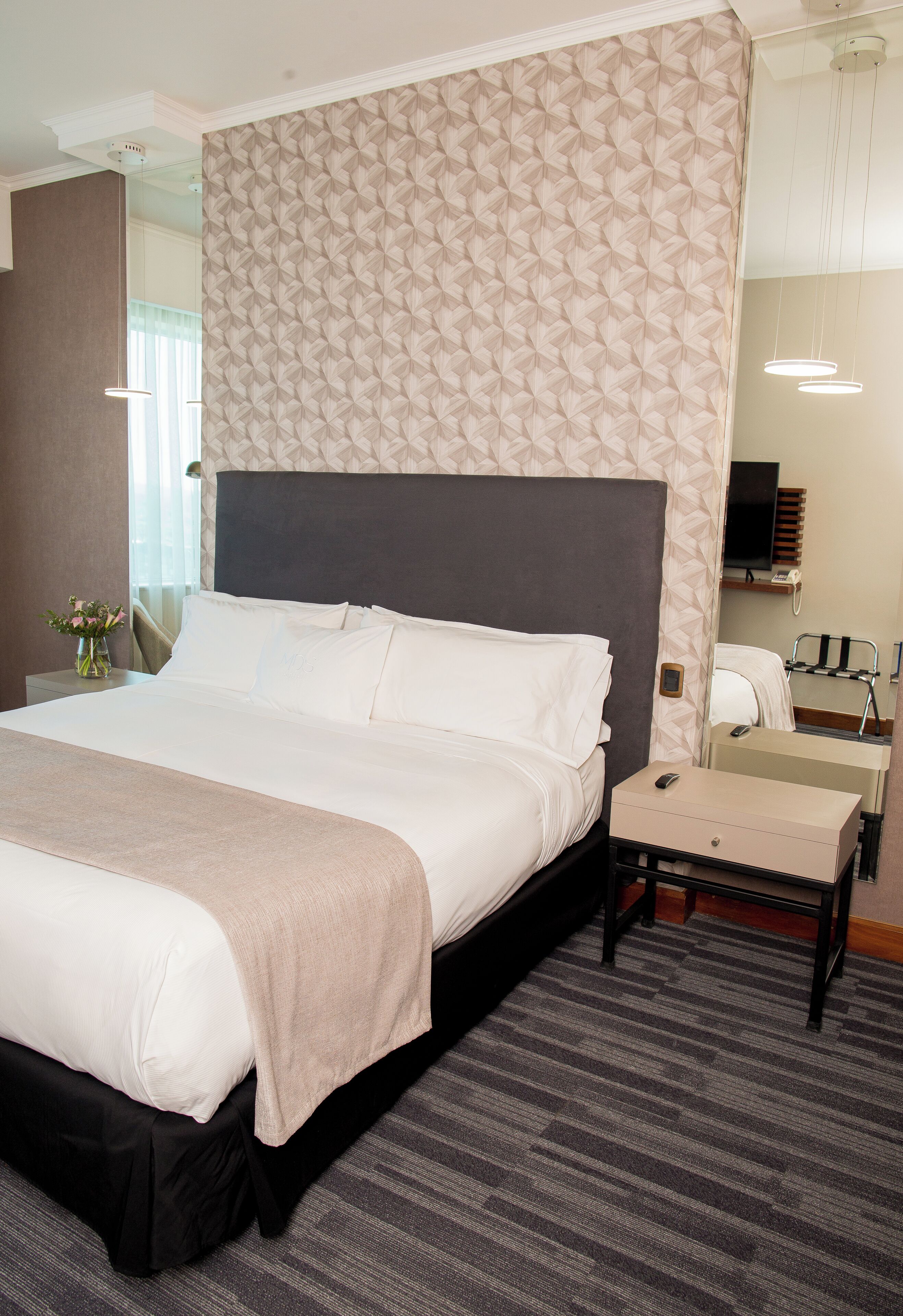 A hotel room with a large bed, a bedside table, a mirror, and a desk with a chair.