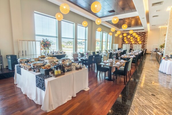 A spacious dining area with multiple tables set for a meal, large windows allowing natural light, and a modern ceiling design with hanging spherical lights.