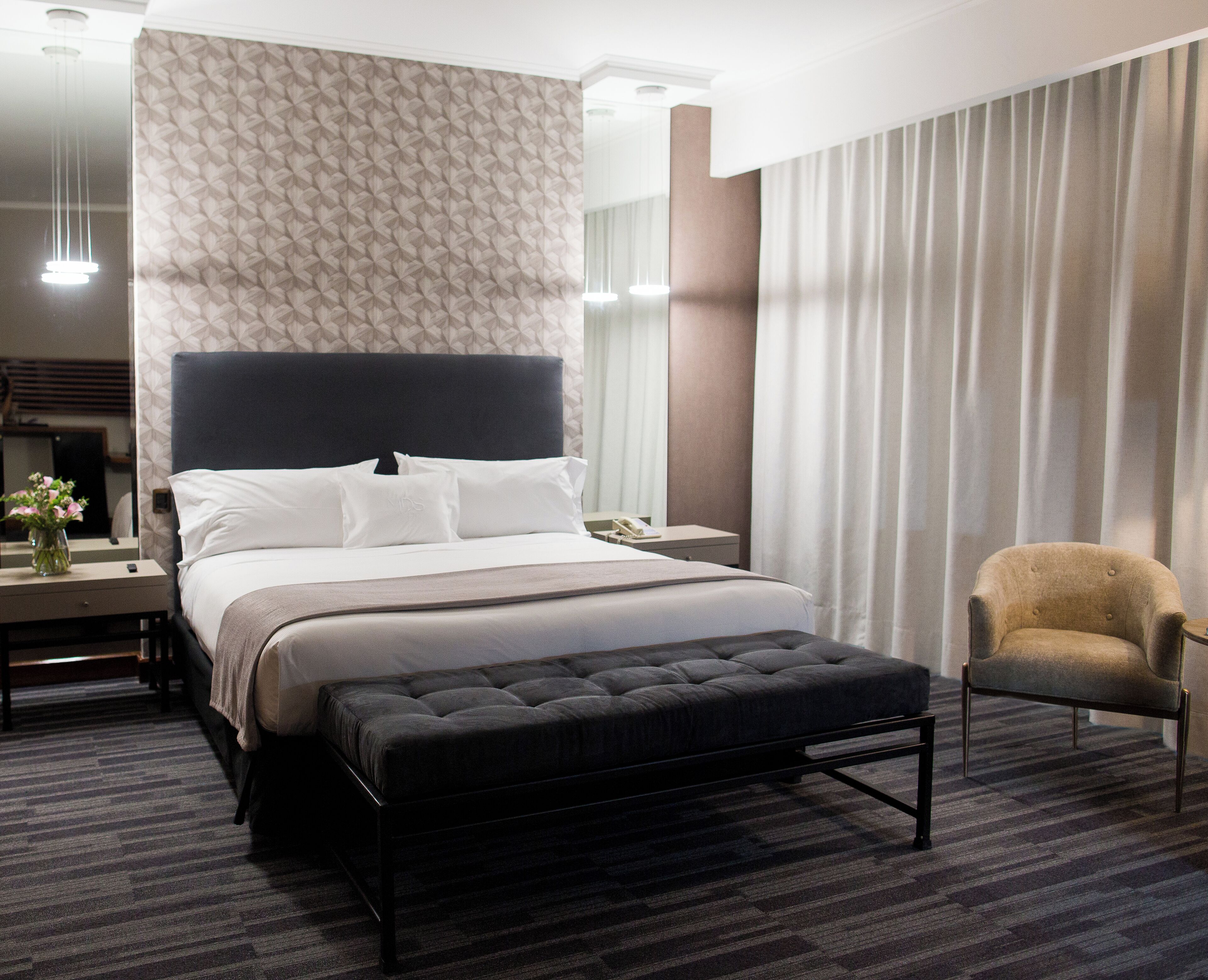 A hotel room with a large bed, a bedside table with a vase of flowers, a chair, and a large window with curtains.