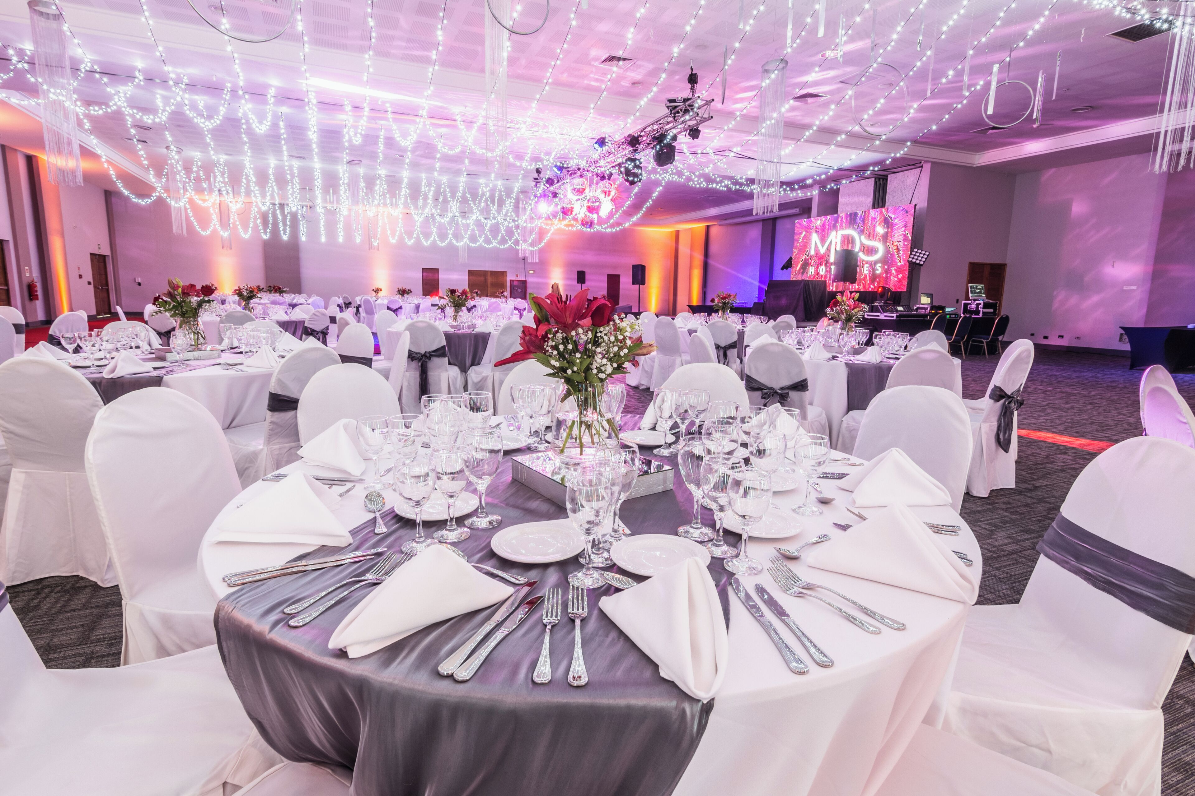 Een banketzaal met ronde tafels, gedekt voor een formeel evenement, met witte tafellakens, bestek en bloemstukken als decoratie.
