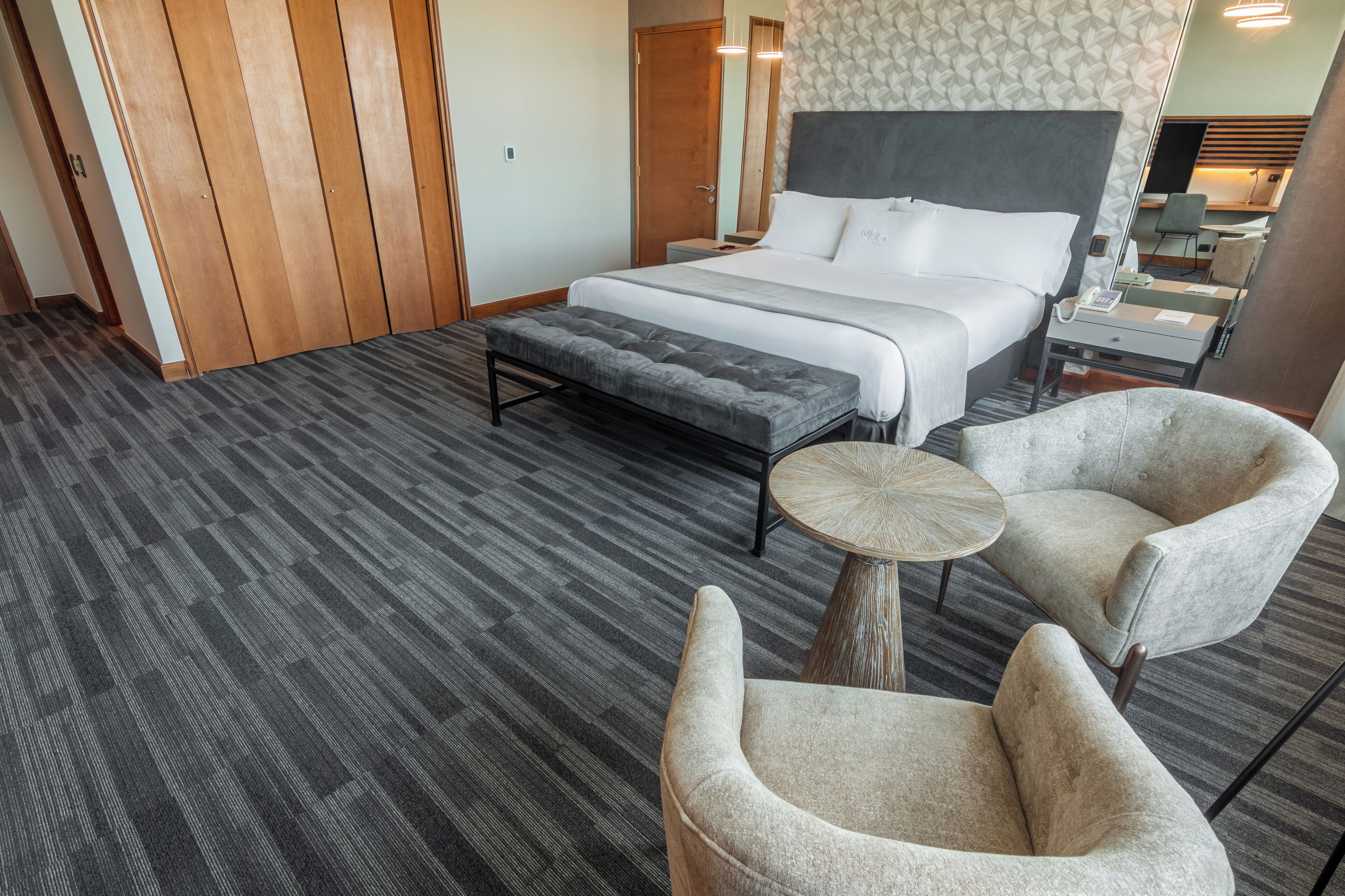 A hotel room with a large bed, a bench, two armchairs, a small round table, and a nightstand with a lamp.