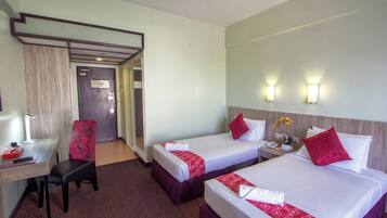 A hotel room with two beds, a desk, a chair, and a small table with a lamp.