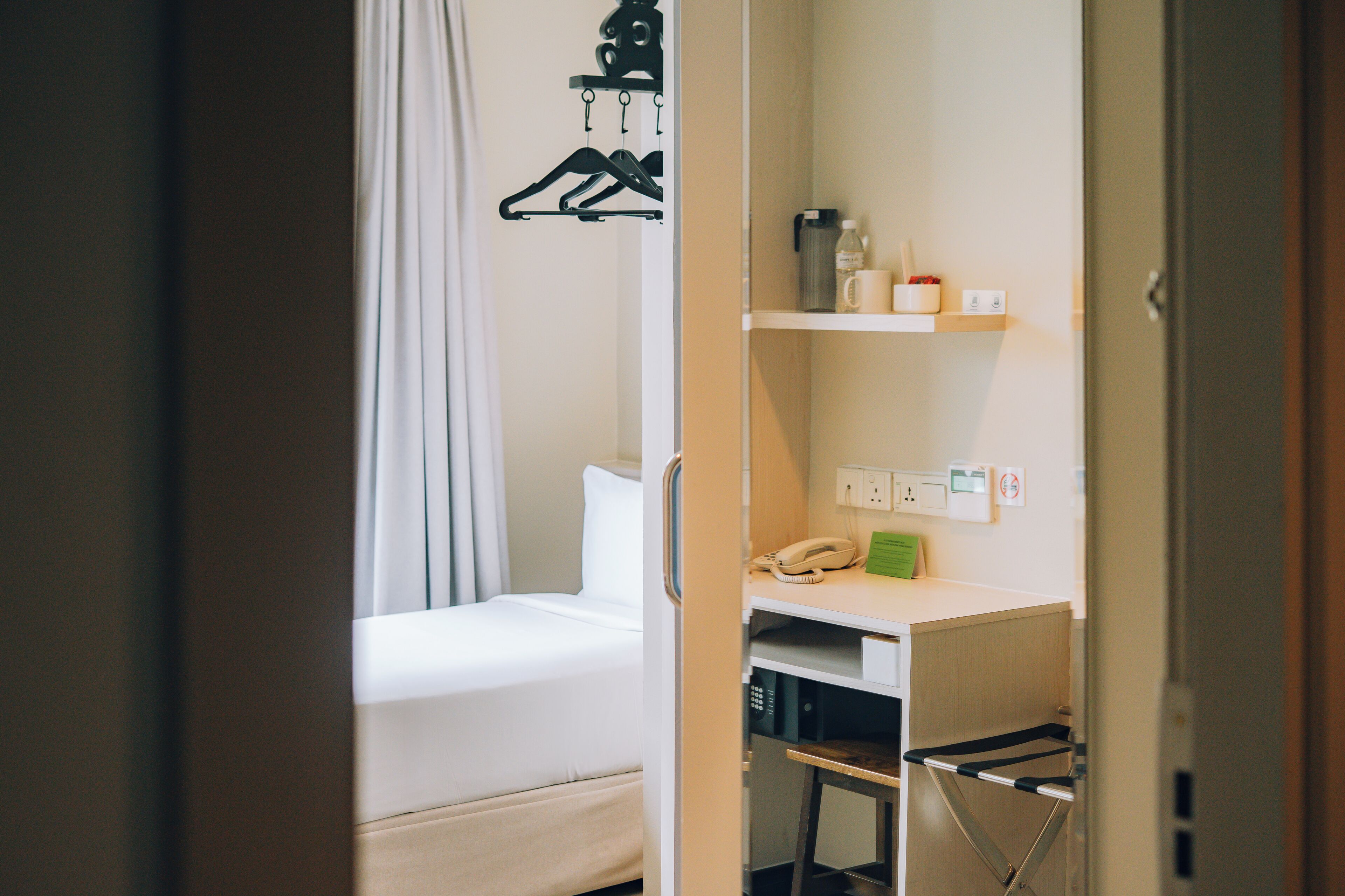 A hotel room with a bed, a desk, a chair, a hanging coat rack, and a small shelf with toiletries.