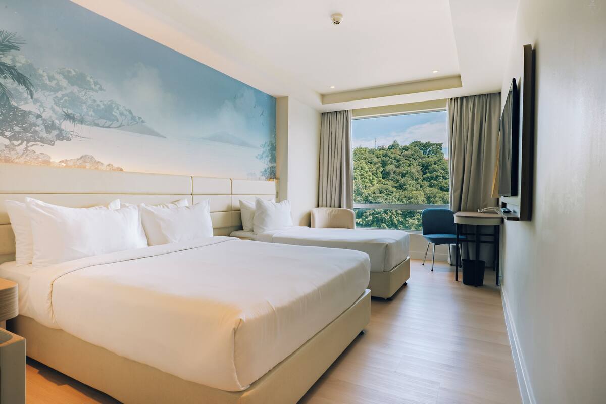 Ein Hotelzimmer mit zwei Betten, einem Schreibtisch, einem Stuhl, einem Fernseher und einem großen Fenster mit Blick auf Grünflächen.