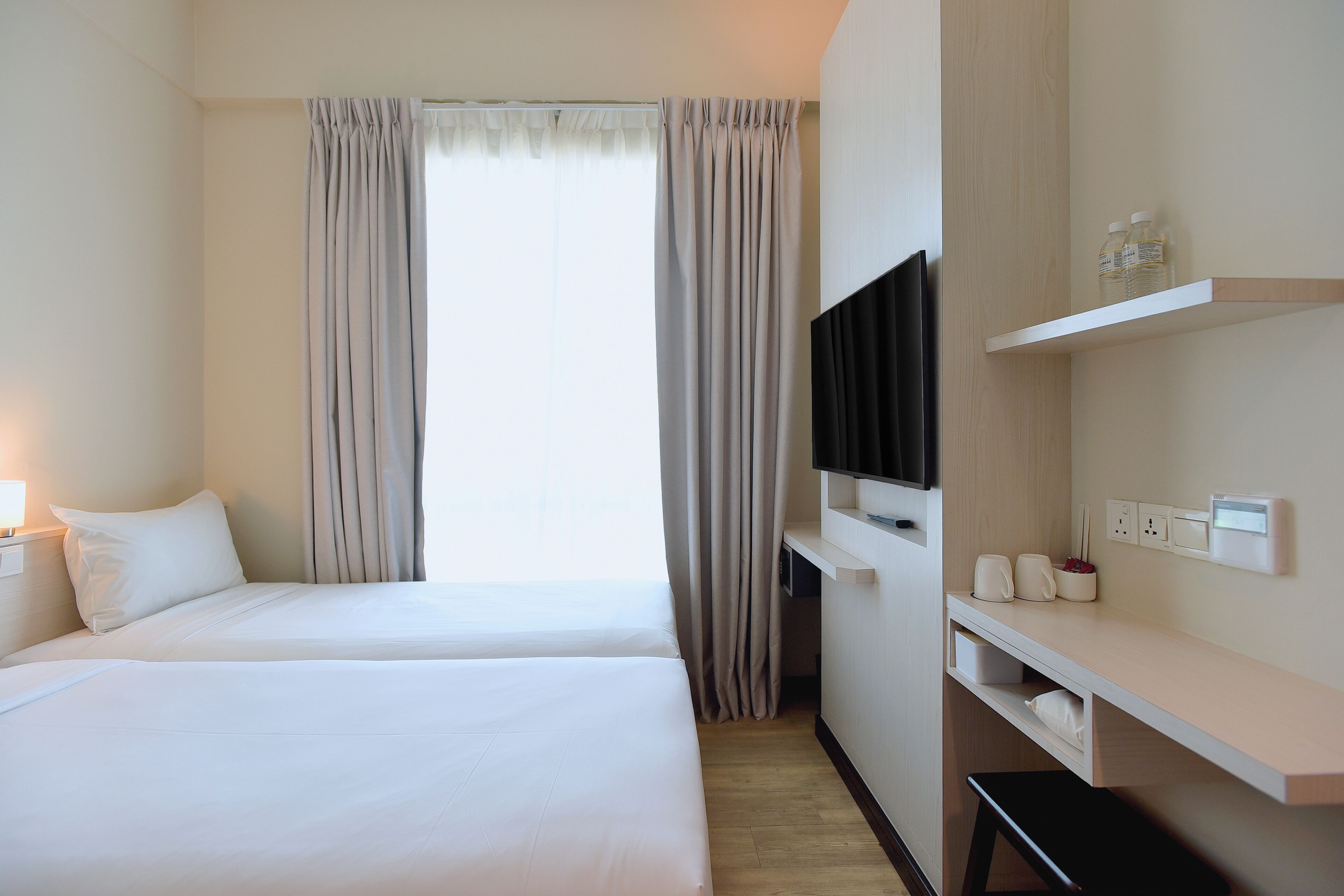 A hotel room with two beds, a TV, a shelf with bottles, and a desk.