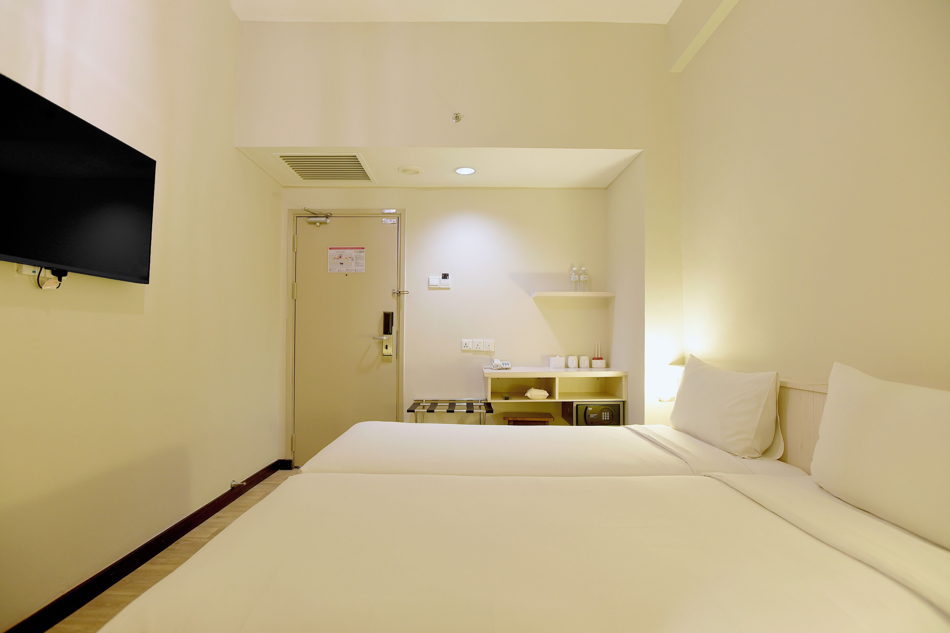 Una habitación de hotel con una cama, un televisor montado en la pared, una mesita de noche y un estante con artículos de aseo.