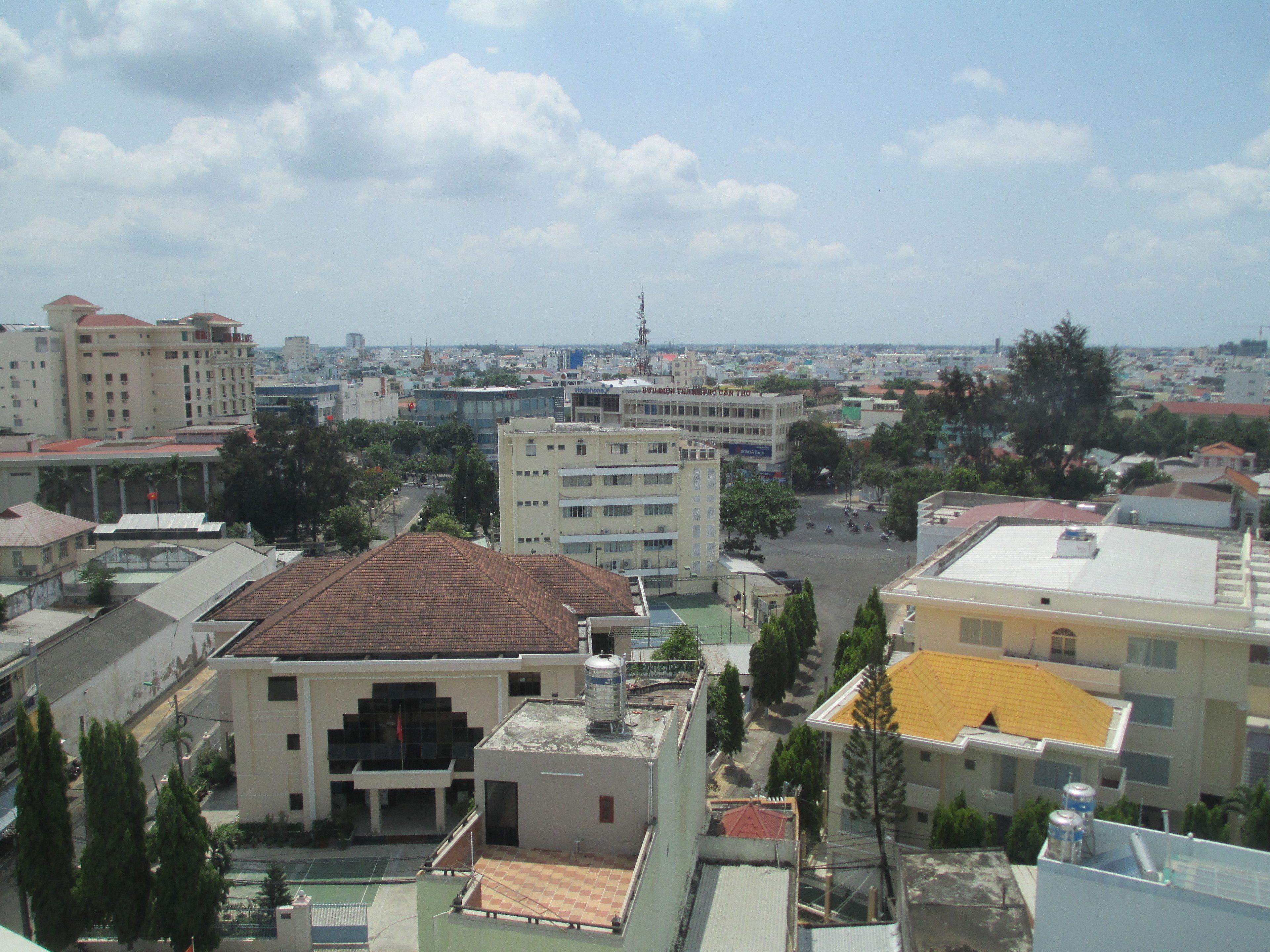 A cityscape with multiple buildings, including a prominent white building with a red roof and a yellow building with a yellow roof. There are trees and a clear sky with clouds.