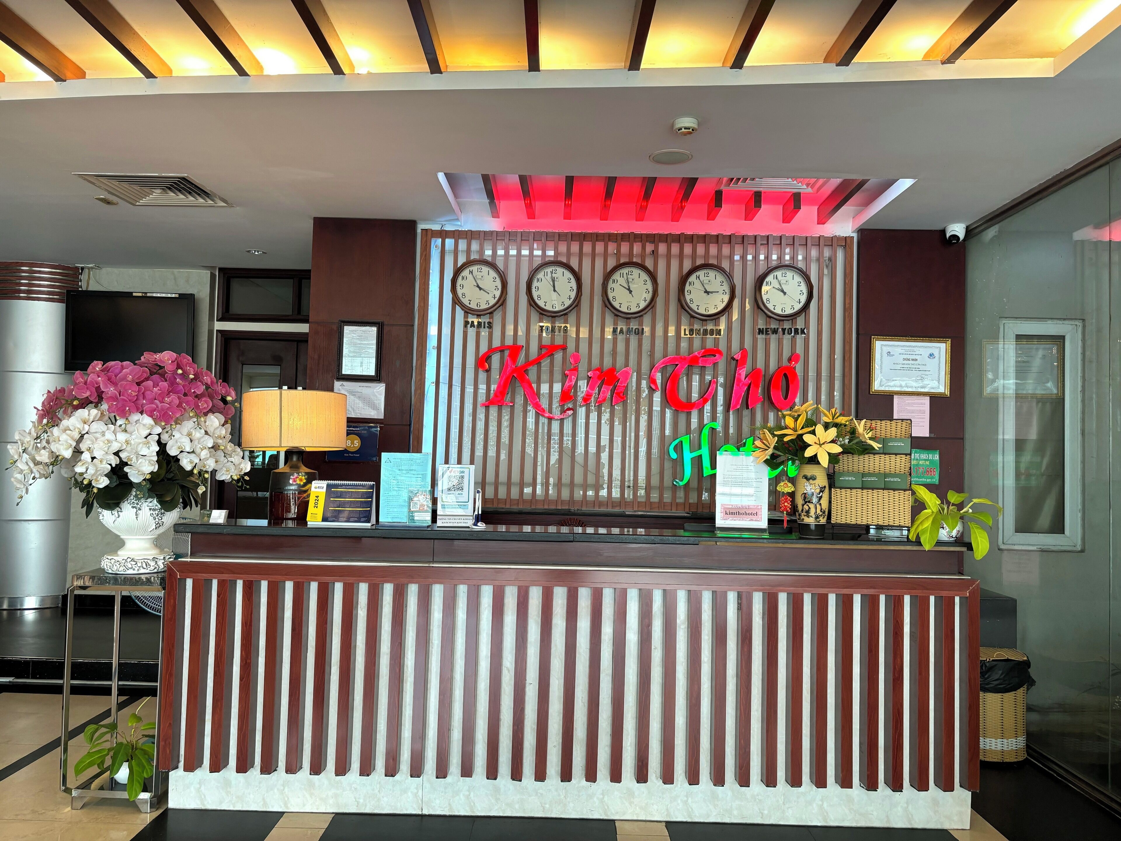 A hotel reception desk with clocks displaying different cities, a large floral arrangement, and a sign reading "Kim Chon Hotel".