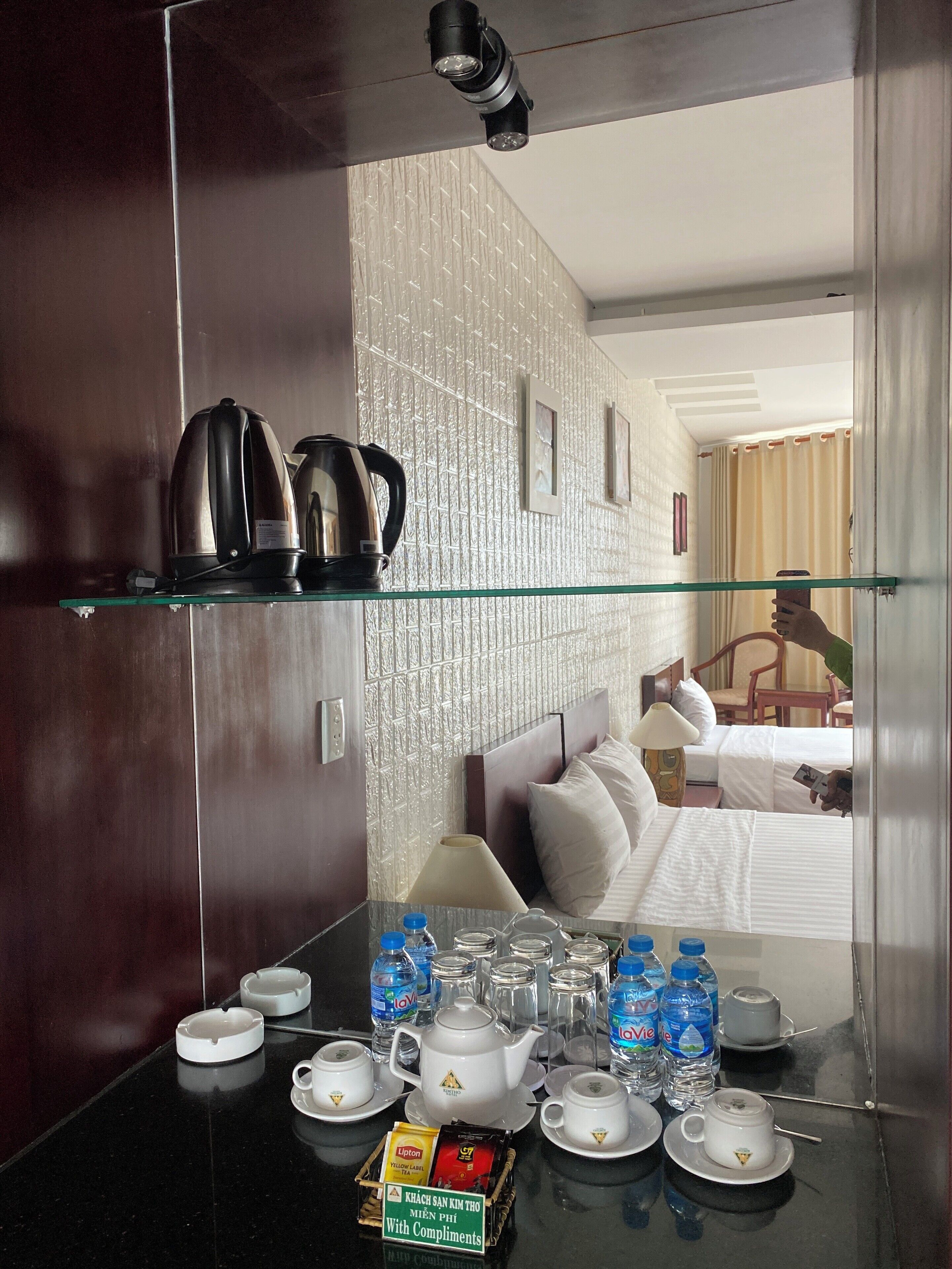 A hotel room with a glass-top table displaying tea sets, water bottles, and a tray of snacks.