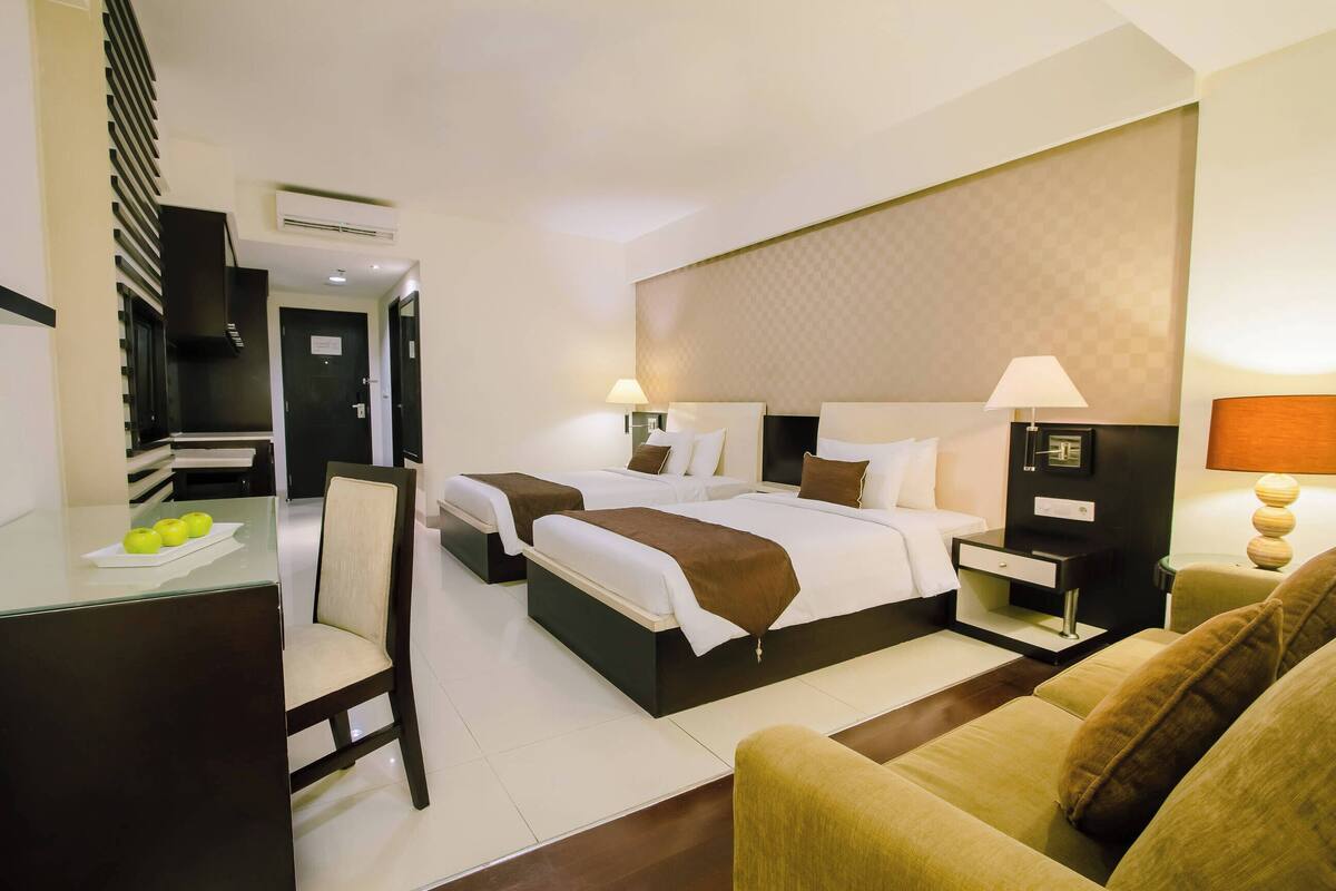 A hotel room with two beds, a desk with apples, a chair, a sofa, and a lamp.