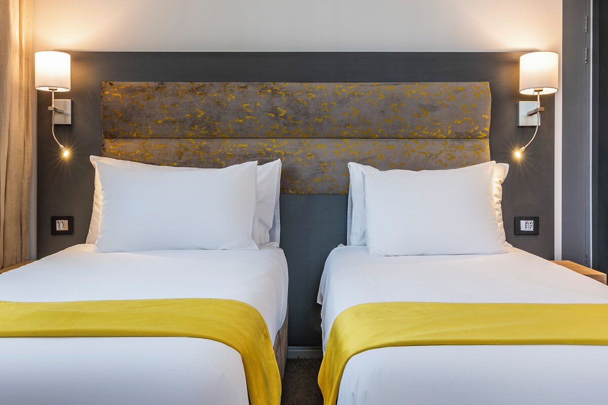 A hotel room with two single beds, each with white bedding and a yellow throw blanket. The headboard has a textured design, and there are wall-mounted lights on either side.