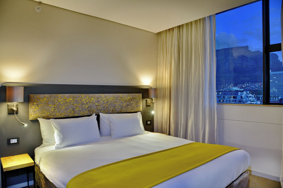 A hotel room with a large bed, a nightstand, a wall-mounted lamp, and a window with a city view.