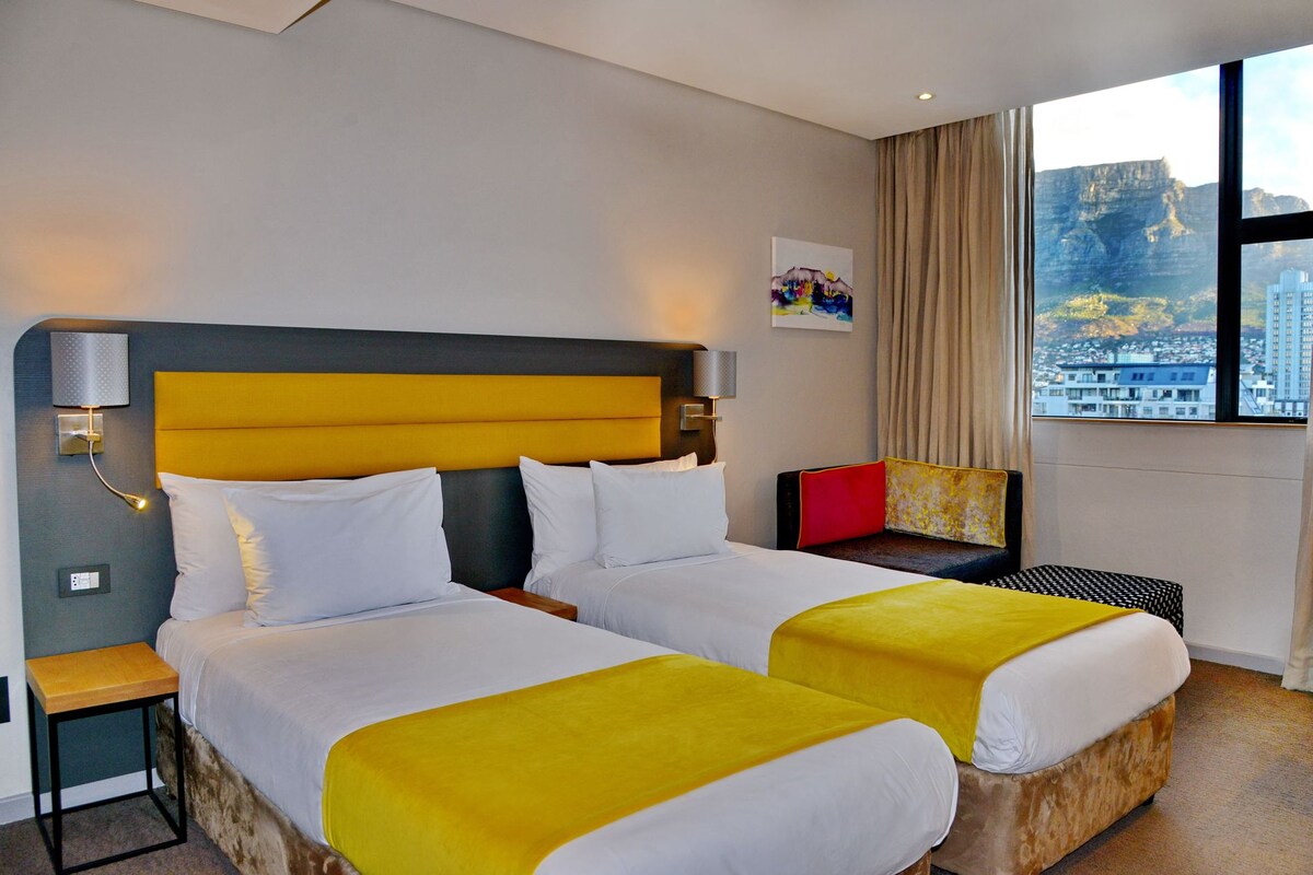 A hotel room with a large bed, two bedside tables, a window with a city view, and a painting on the wall.