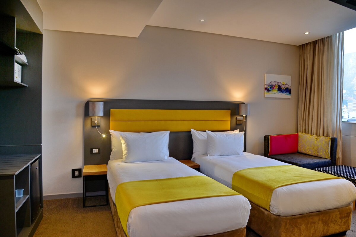 A hotel room with two beds, a bedside table, a chair, a painting, and a window with curtains.