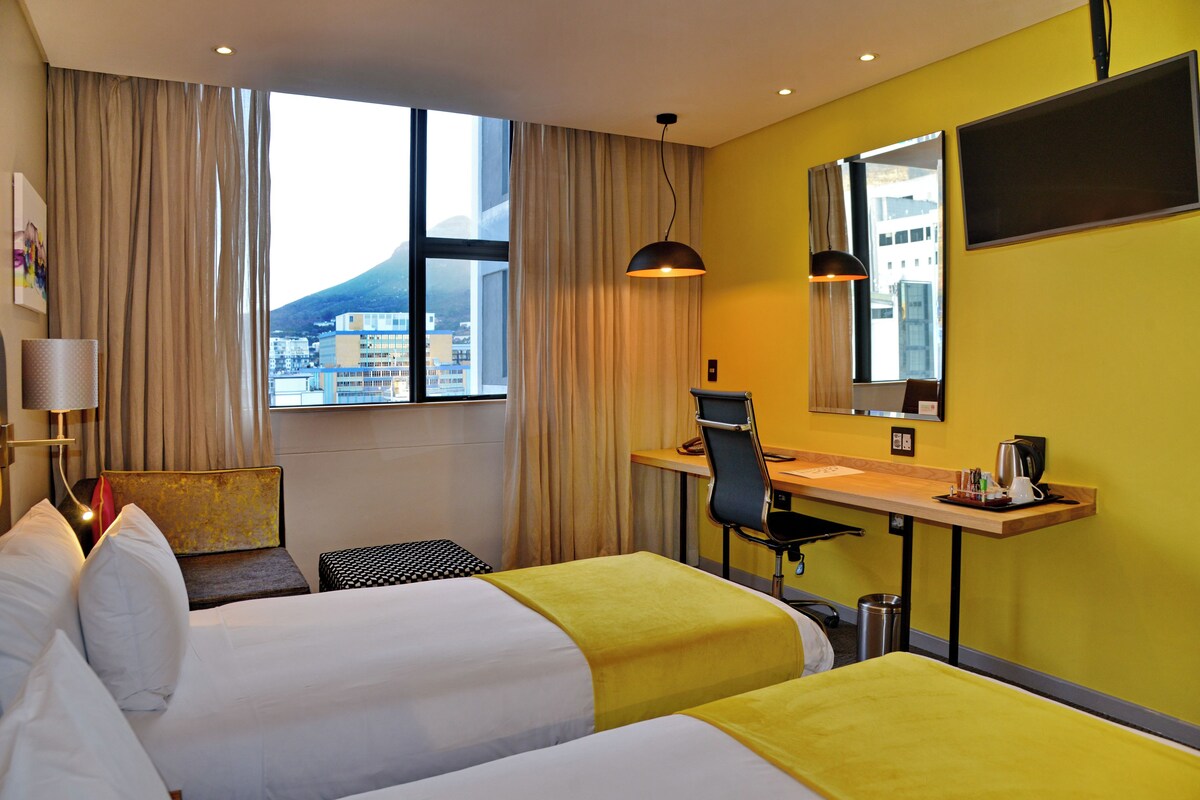 A hotel room with two beds, a desk, a chair, a lamp, a trash can, and a window with a view of a mountain.