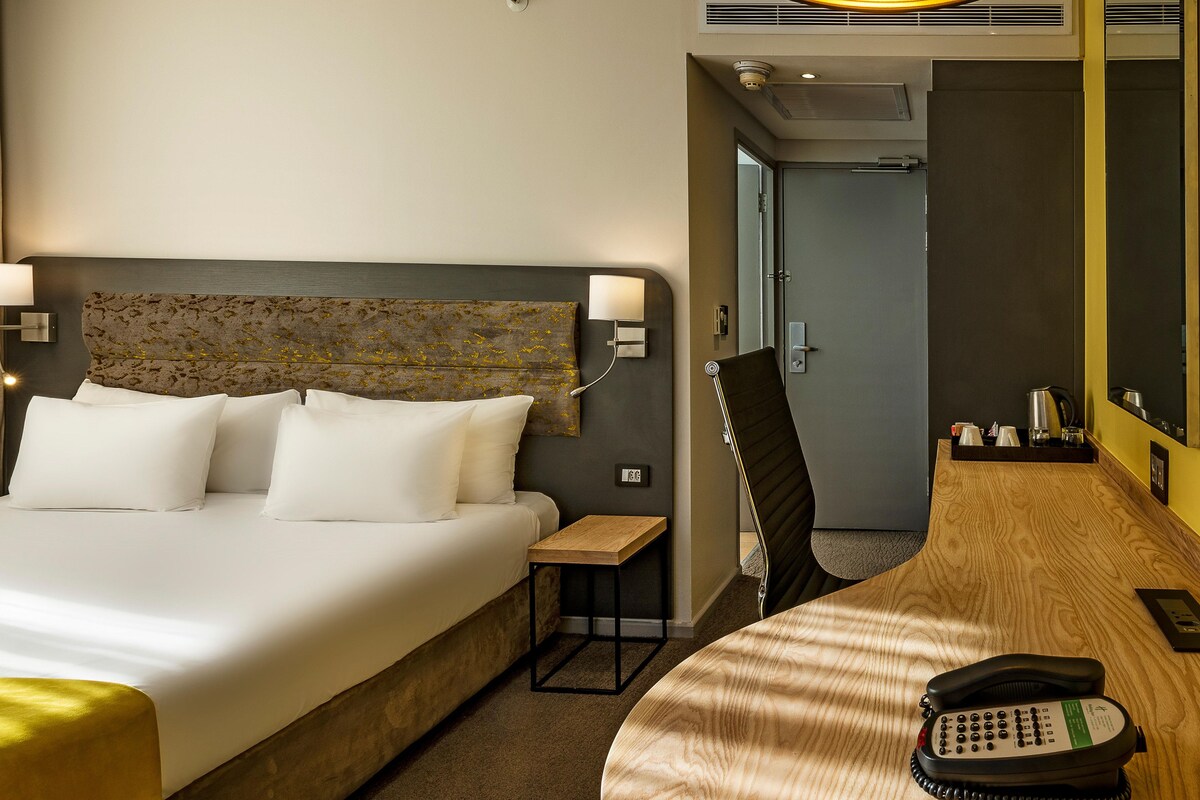 A hotel room with a bed, bedside table, lamp, chair, desk, phone, and a mirror.