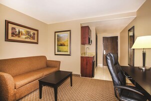 A hotel room with a sofa, a small table, a chair, a desk, and a lamp. There are framed pictures on the wall and a door leading to the bathroom.