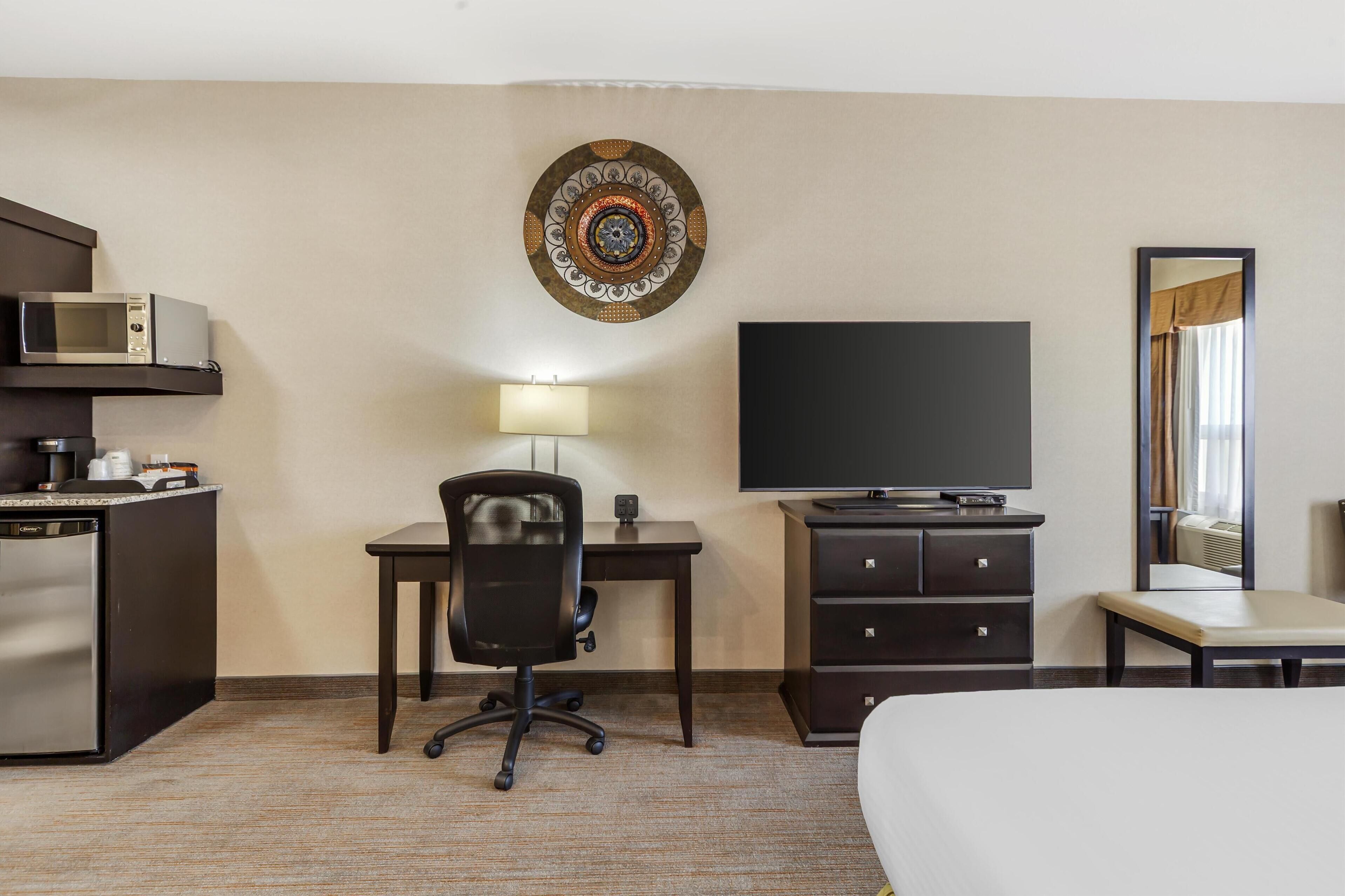 A hotel room with a bed, a desk with a chair, a flat-screen TV, a microwave, a mini-fridge, a dresser, and a decorative wall piece.