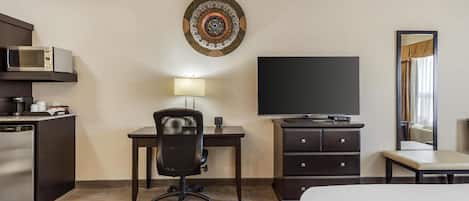 Habitación de hotel con cama, escritorio y silla, televisor de pantalla plana, microondas, refrigerador pequeño, cómoda y un adorno en la pared.