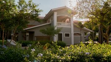 A two-story house with a balcony, surrounded by lush greenery and flowering plants.