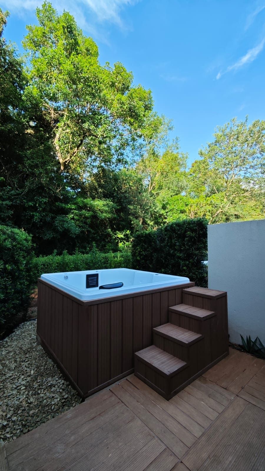 A hot tub with wooden steps leading up to it, surrounded by greenery and a clear blue sky.
