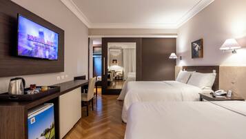 Quarto de hotel com televisão exibindo 'Dreamland Museum of Cinema', uma mesa com chaleira, mini-geladeira e uma cadeira. Há duas camas com roupa branca, uma mesa de cabeceira com telefone e um abajur na parede.