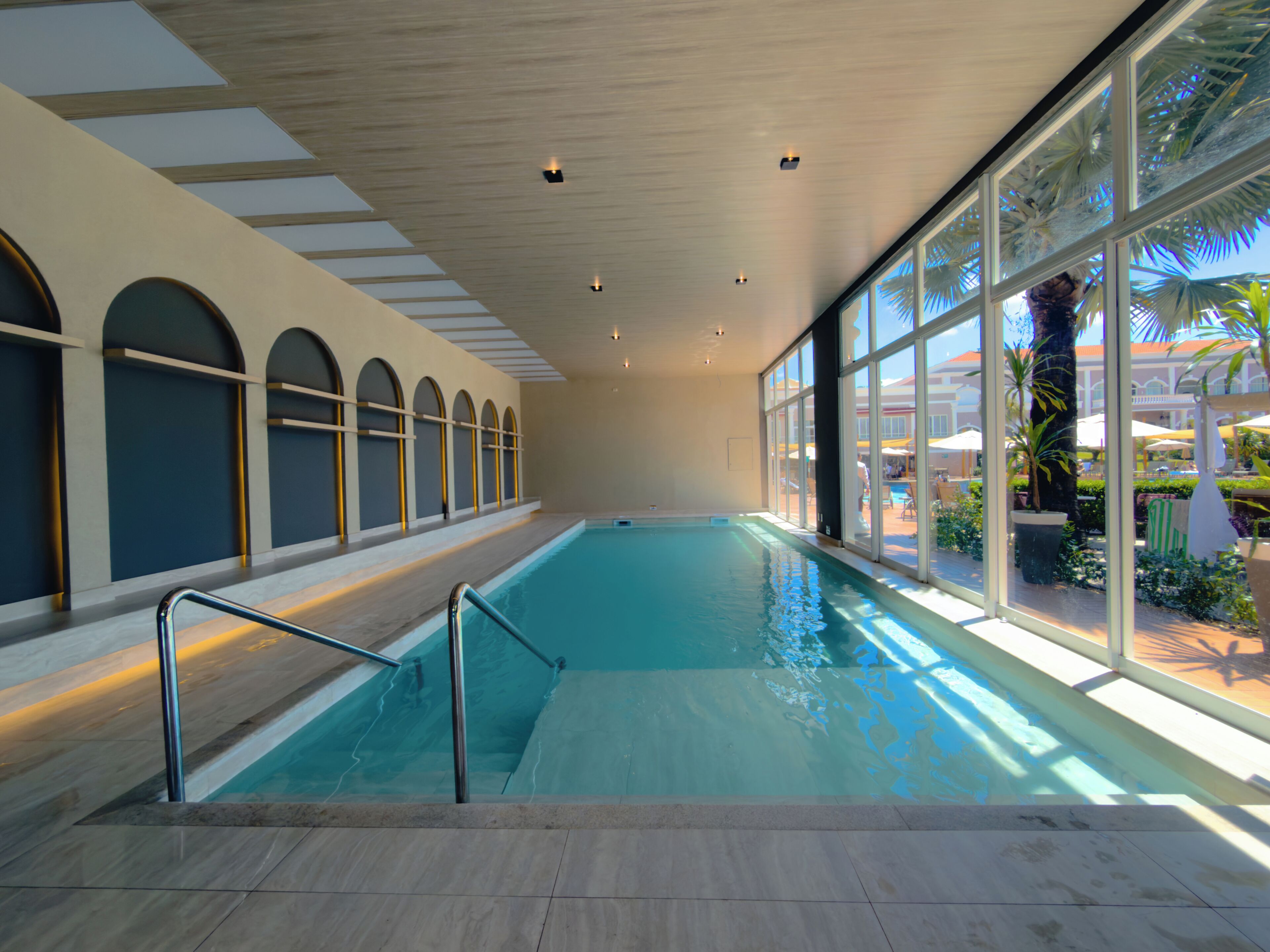 Una piscina interior con paredes de vidrio transparente, suelo de baldosas e iluminación empotrada en el techo.
