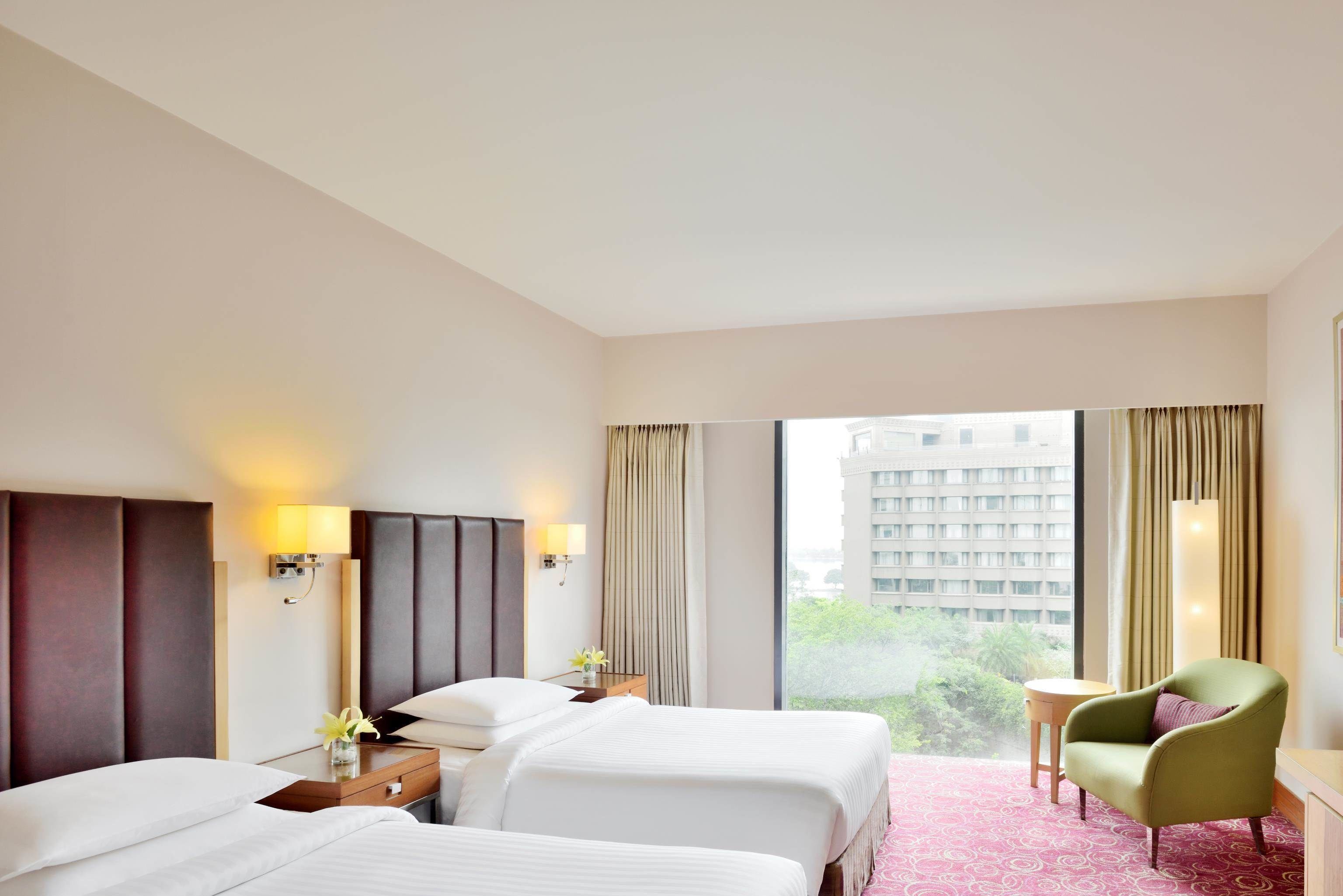 A hotel room with two beds, a small table, a chair, and a view of a building and greenery outside.