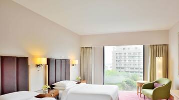 Habitación de hotel con dos camas, una mesita, una silla y vistas a un edificio y zonas verdes.