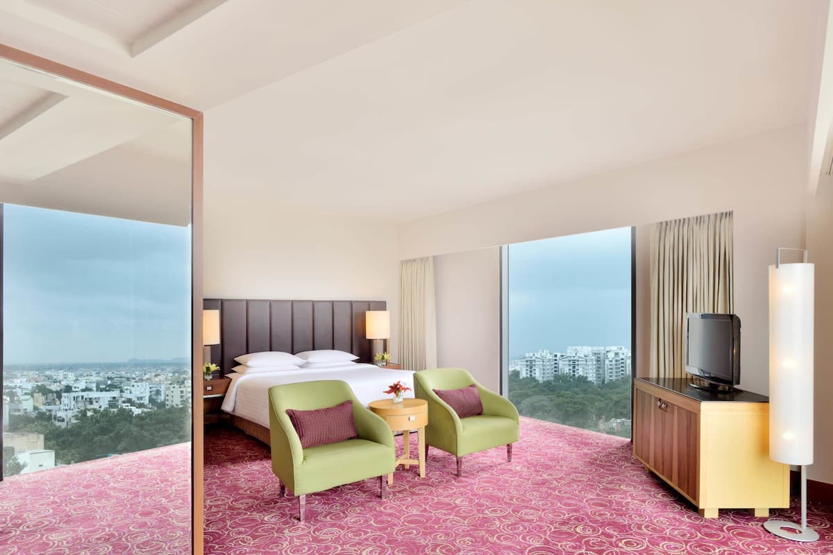 A hotel room with a bed, two armchairs, a TV, a small table, and a large window offering a city view.