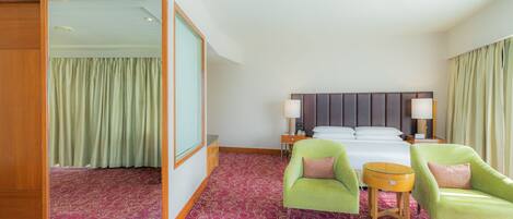 A hotel room with a large bed, two green armchairs, a small round table, and a mirror.