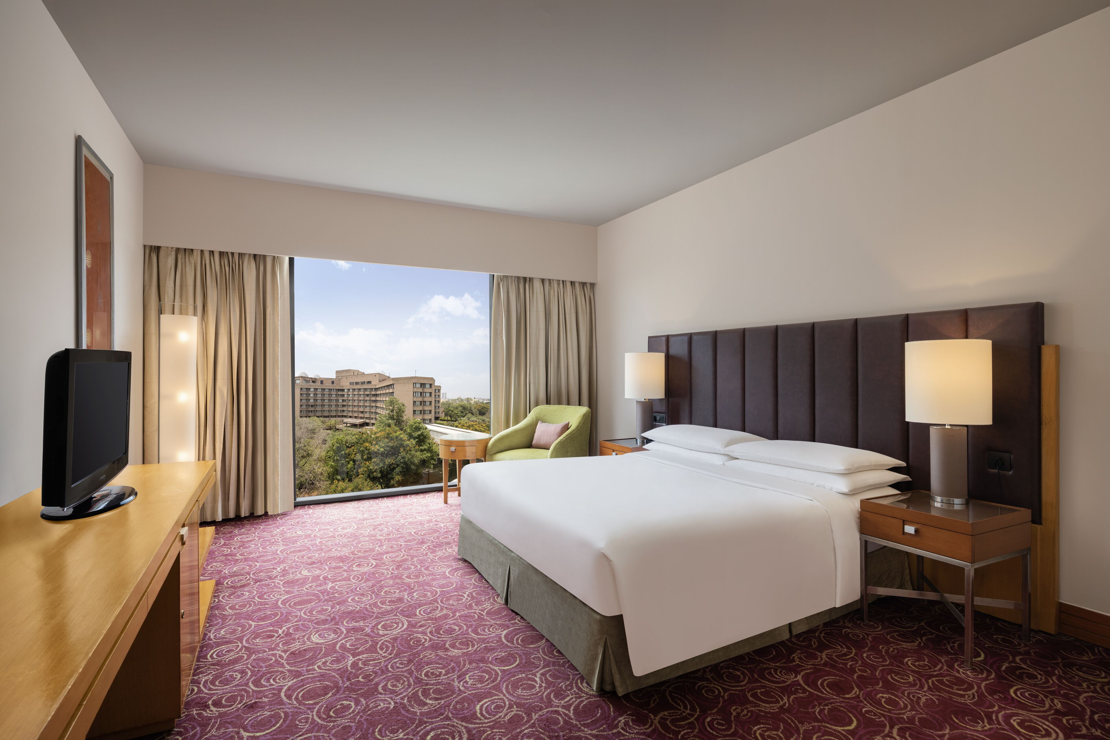 A hotel room with a large bed, a desk with a TV, a chair, and a nightstand with a lamp.