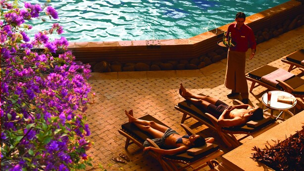Un espace piscine agrémenté d’une cascade, de chaises longues et d’une personne se tenant près de la piscine.