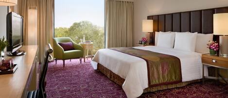 Habitación de hotel con una cama grande, un escritorio con televisor, una silla y vistas a árboles por la ventana.