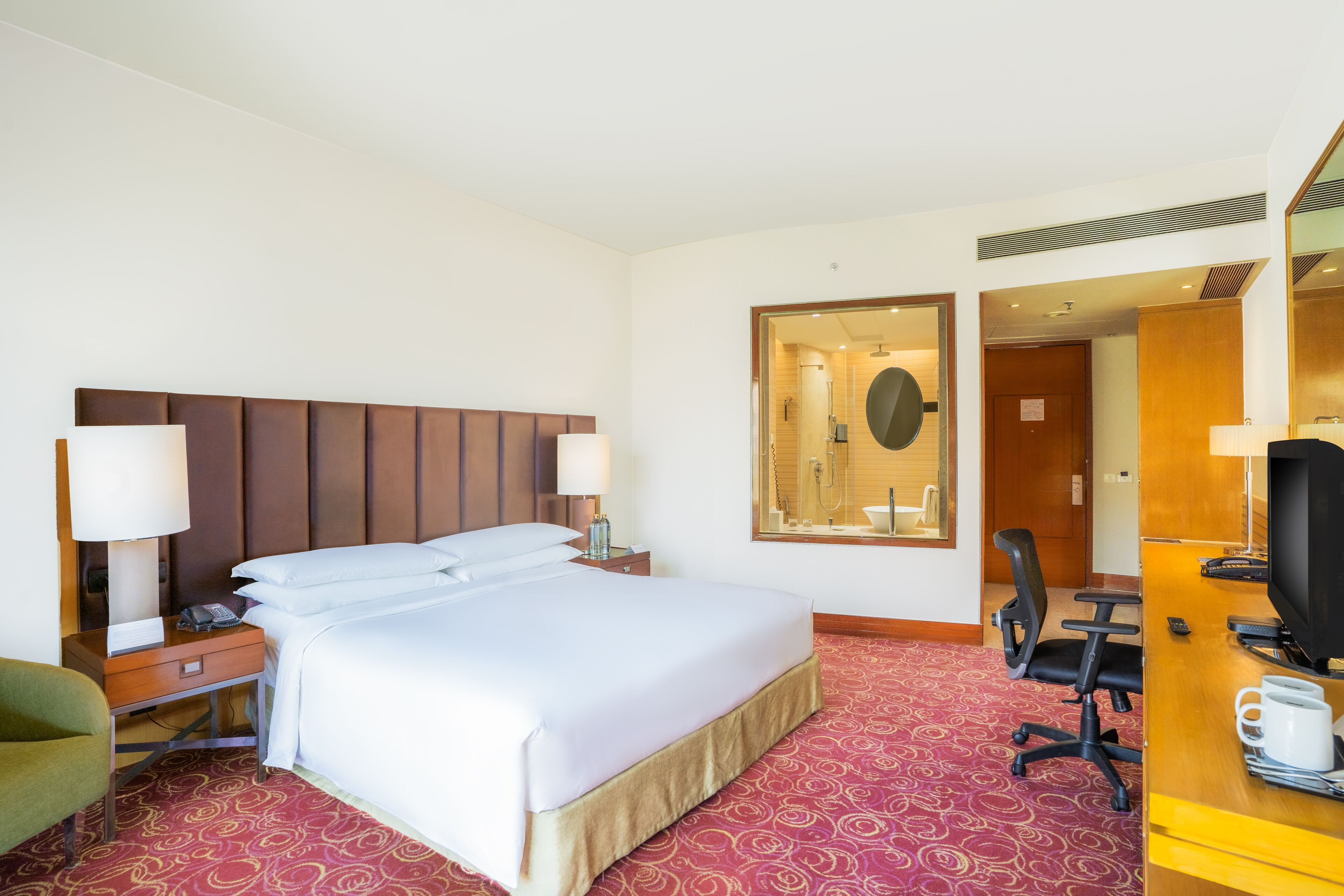 Quarto de hotel com uma cama grande, mesas de cabeceira com luminárias, uma escrivaninha com cadeira, uma televisão e um espelho.